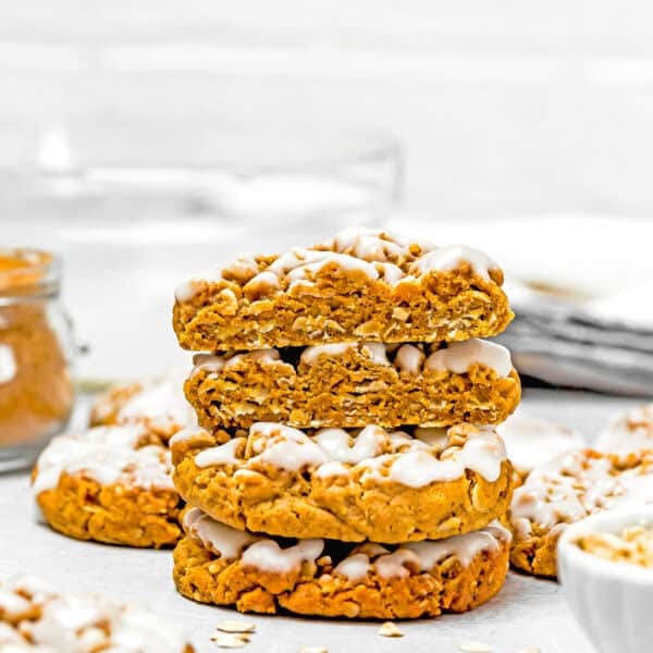 A stack of glazed, eggless iced gingerbread oatmeal cookies on a white surface, surrounded by additional cookies and oats in the background.