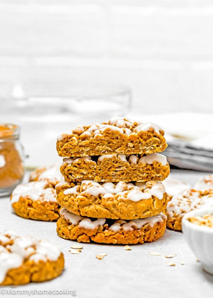 A stack of glazed, eggless iced gingerbread oatmeal cookies on a white surface, surrounded by additional cookies and oats in the background.