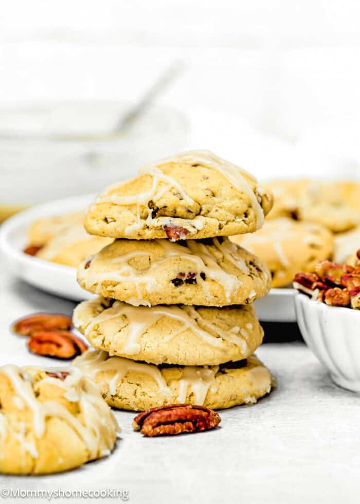 A stack of four iced eggless maple brown sugar cookies with pecans on a light surface. A few pecans and more cookies on a plate are visible in the background.