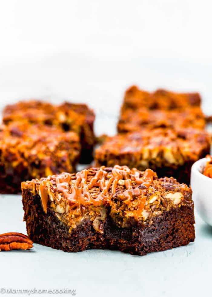 Close-up of eggless pecan pie brownies drizzled with caramel. Several brownies are stacked in the background, and a small bowl of pecans is visible on the side.