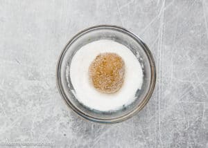 A round ball of dough, reminiscent of eggless gingersnap cookies, is coated in sugar and rests in a small glass bowl on a textured gray surface.