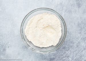 A glass bowl filled with flour sits on a textured gray surface, ready for crafting delightful eggless gingersnaps cookies.