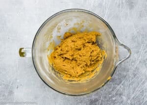 A glass mixing bowl brimming with eggless gingersnaps cookie dough sits on a scratched, metallic surface.