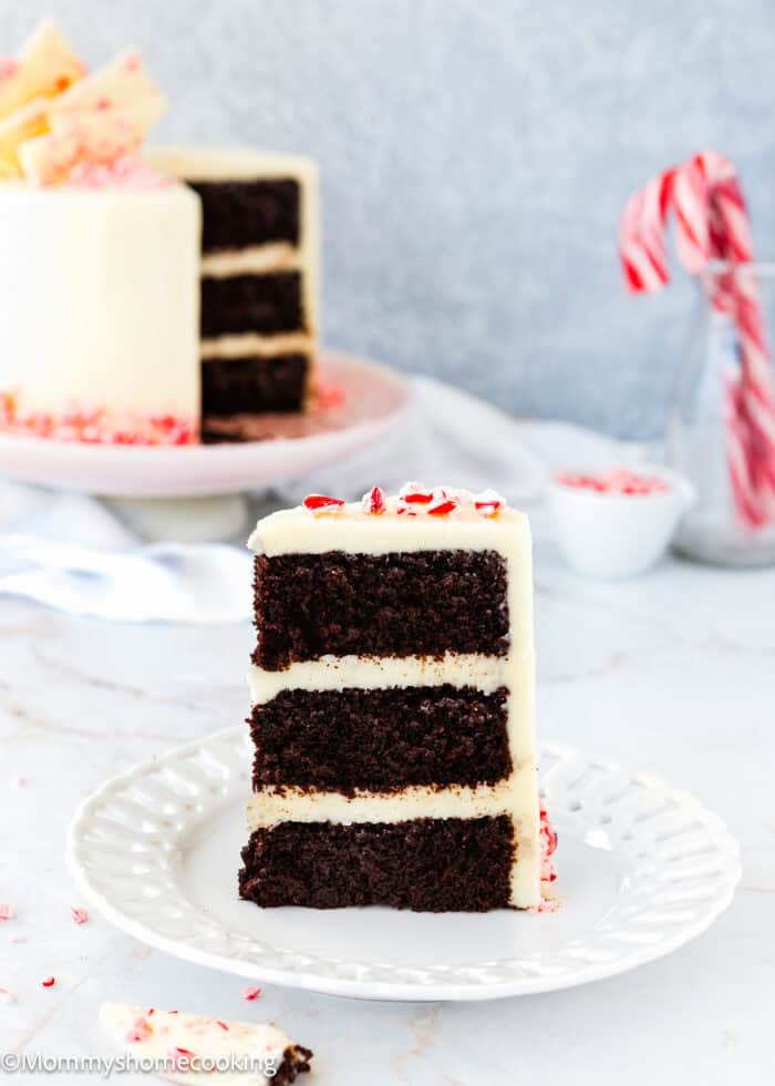 A slice of eggless chocolate peppermint layer cake with white frosting and crushed candy canes sits elegantly on a white plate. In the background, the whole cake and candy canes create a festive atmosphere.