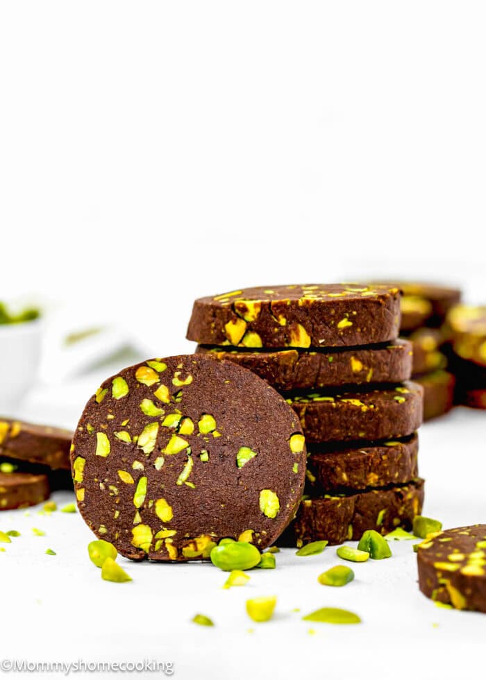 A stack of eggless chocolate pistachio slice and bake cookies sits elegantly on a white background, surrounded by scattered pistachios.