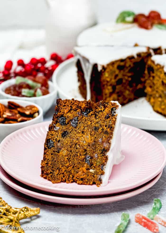 An eggless Christmas fruit cake with white icing sits on a pink plate, surrounded by an array of fruits and nuts.