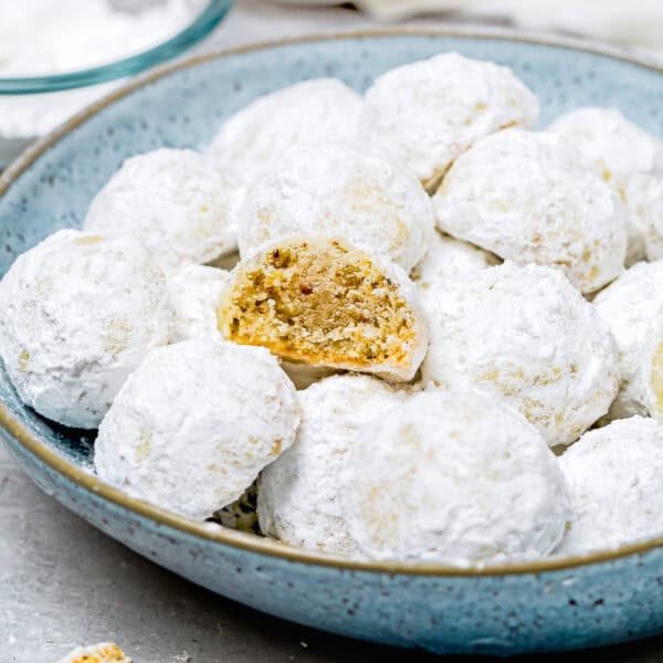 A blue plate filled with eggless snowball cookies, topped with delicate powdered sugar. One cookie is partially eaten, revealing a nutty interior.