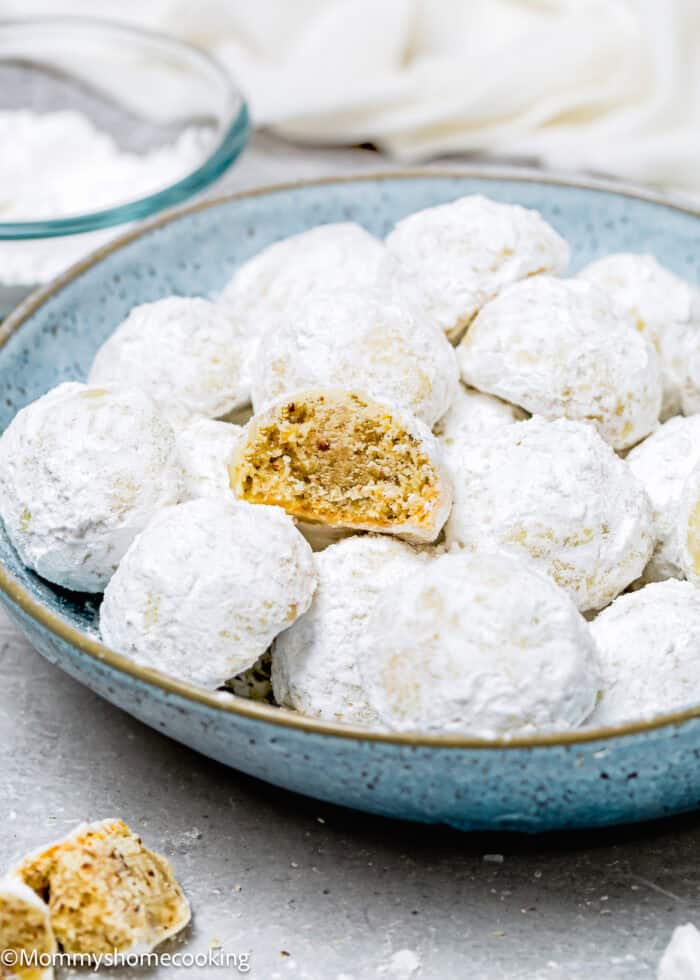 A blue plate filled with eggless snowball cookies, topped with delicate powdered sugar. One cookie is partially eaten, revealing a nutty interior.