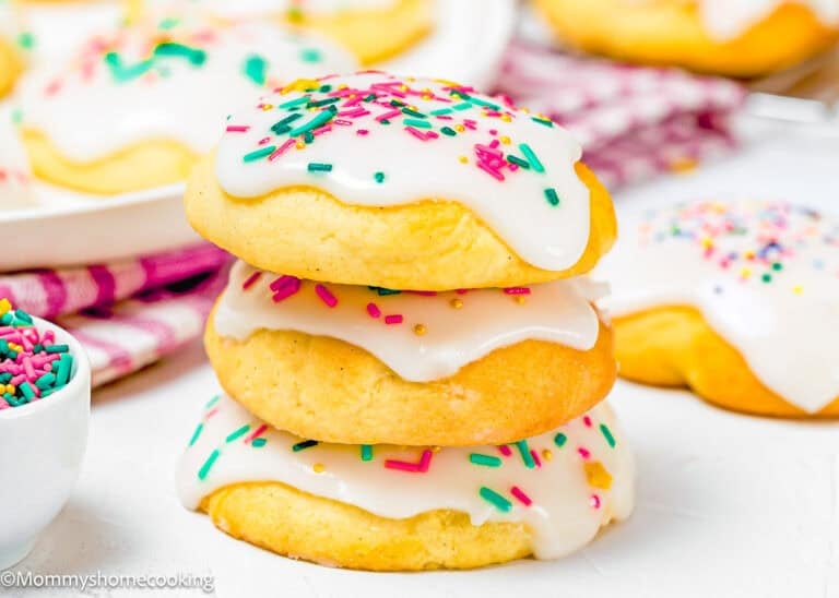 Three round Eggless Italian Ricotta Cookies with white icing and colorful sprinkles are stacked on top of each other, with more cookies and a bowl of sprinkles in the background.