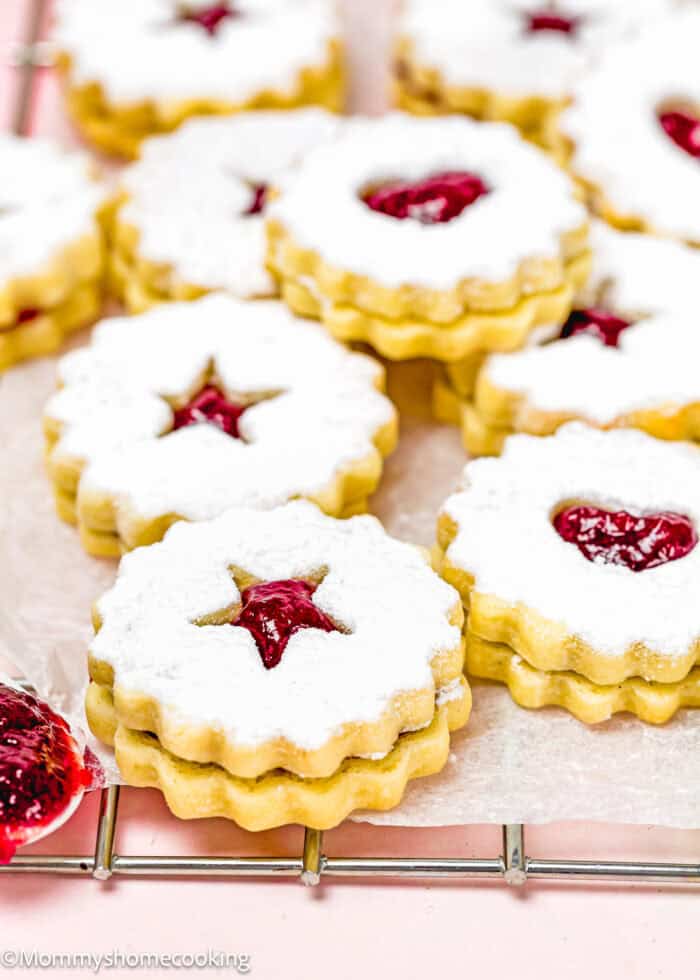 Eggless Linzer Cookies (Nut-Free) with powdered sugar and raspberry filling, featuring star and heart cutouts, are arranged on a cooling rack.