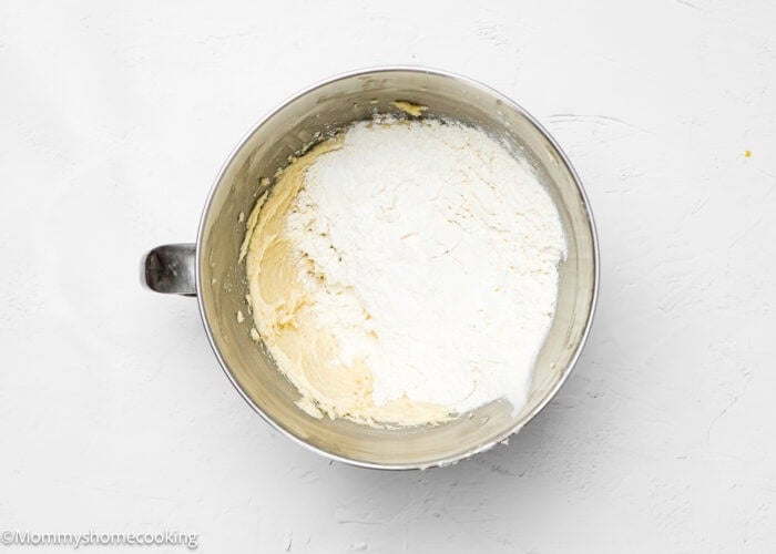 A mixing bowl with partially combined flour and batter on a white surface suggests the beginning of creating eggless lemon slice and bake cookies.