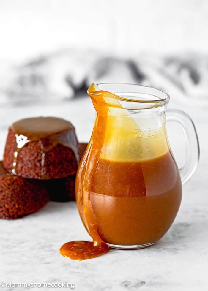 A glass pitcher brimming with caramel sauce, some elegantly spilling over the side, sits next to two delectable chocolate cakes on a marble surface. Perfect for those exploring how to make toffee sauce yourself.