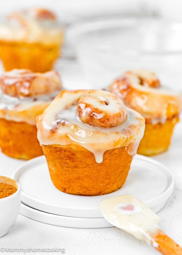 Three glazed cinnamon rolls on small white plates, reminiscent of quick eggless cinnamon roll muffins, with a white spatula and a bowl of cinnamon nearby.