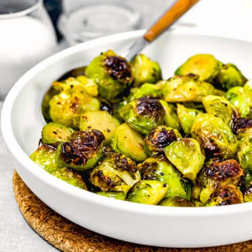 A white bowl brimming with Easy Maple Roasted Brussels Sprouts, some perfectly charred, rests on a cork mat. A wooden-handled spoon nestles among the sprouts, while salt and pepper shakers stand ready in the background.