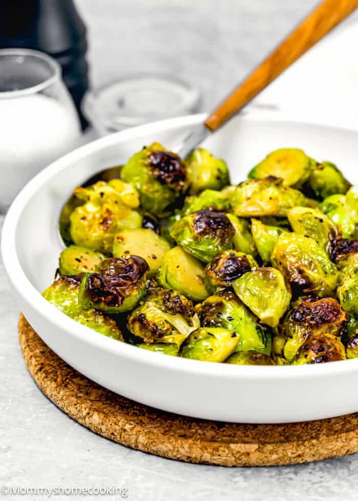 A white bowl brimming with Easy Maple Roasted Brussels Sprouts, some perfectly charred, rests on a cork mat. A wooden-handled spoon nestles among the sprouts, while salt and pepper shakers stand ready in the background.