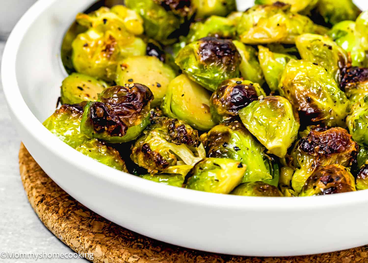 A white bowl filled with easy maple roasted Brussels sprouts, some with browned edges, placed on a cork mat.