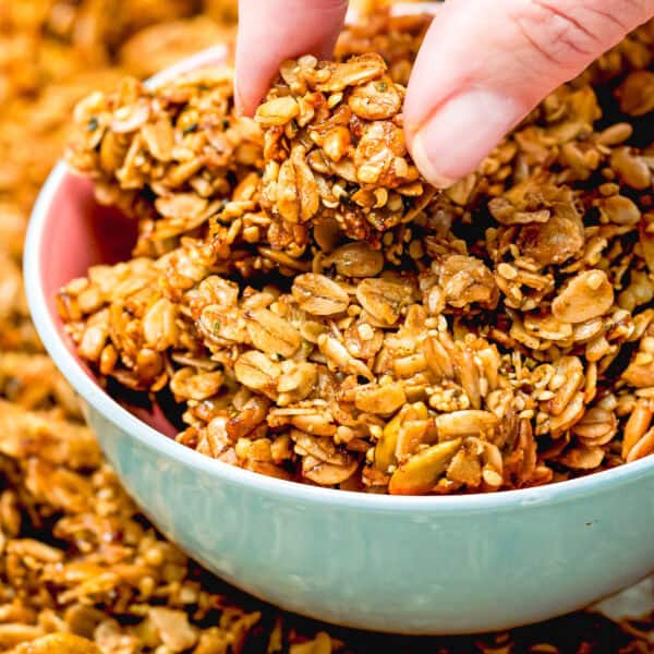 A hand picks up a cluster of nut-free homemade granola from a bowl, with a spread of simple granola pieces in the background.