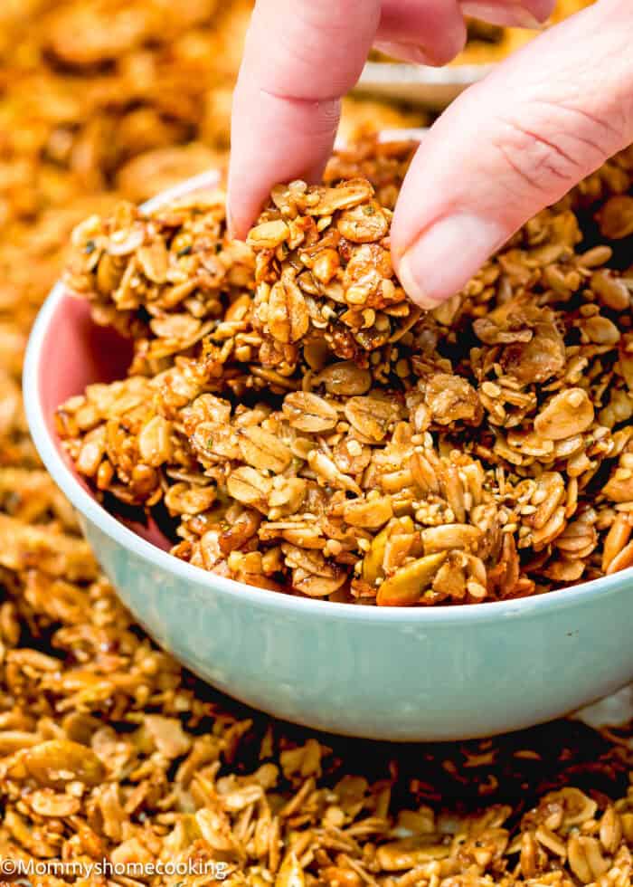 A hand picks up a cluster of nut-free homemade granola from a bowl, with a spread of simple granola pieces in the background.