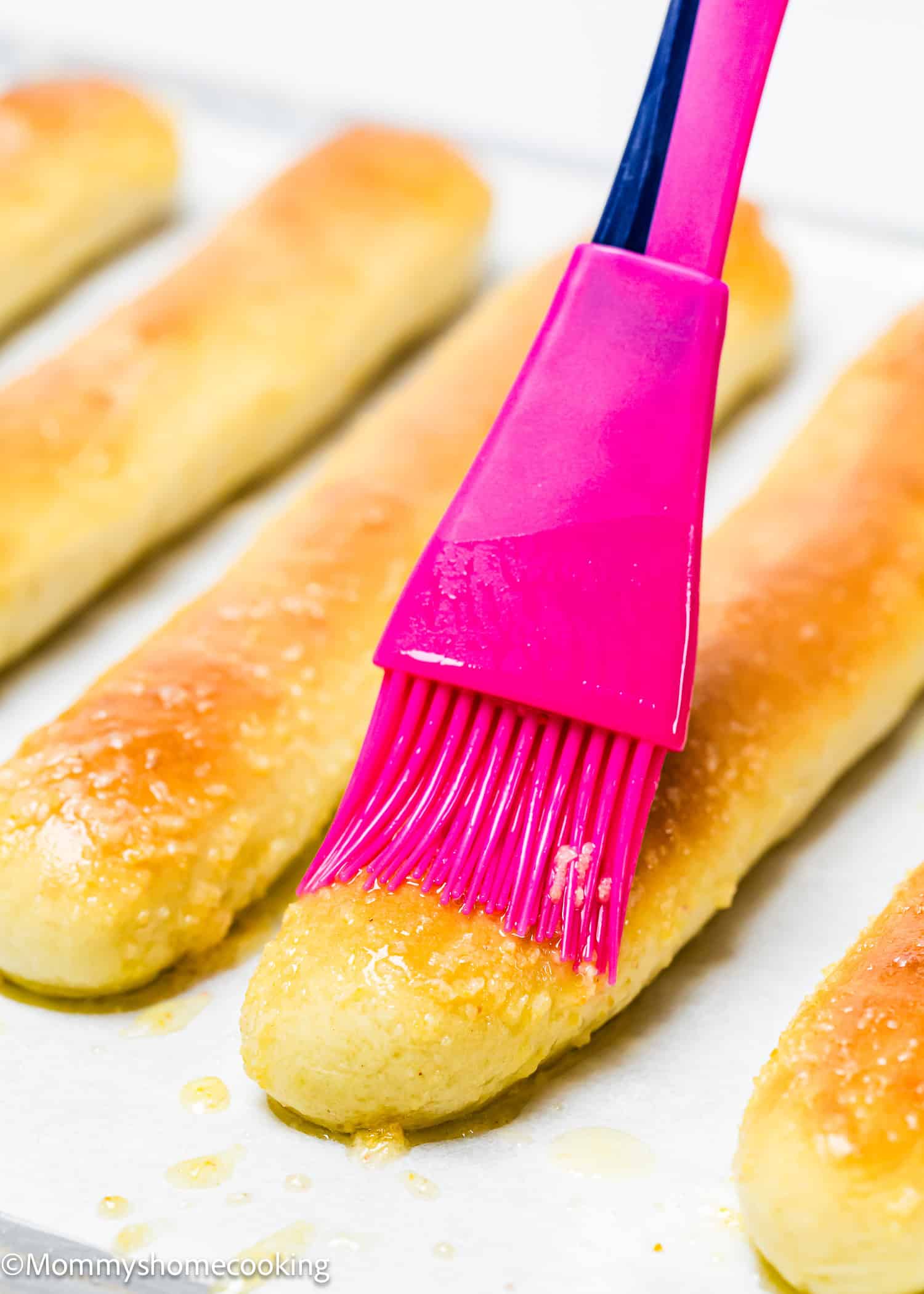 A pink brush smoothly applies butter over quick and easy breadsticks, which are egg-free and dairy-free, lined on parchment paper.