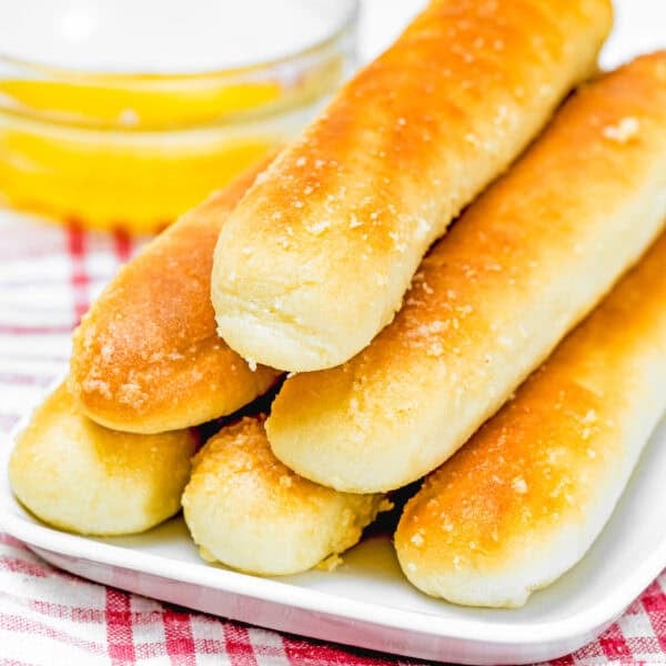 A stack of golden-brown, quick and easy breadsticks (egg-free & dairy-free) rests on a white plate. In the background, a small glass bowl of melted butter complements the scene, all set atop a red and white checkered cloth.