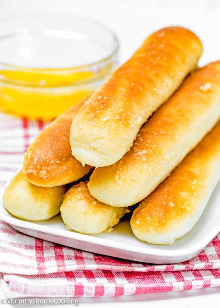 A stack of golden-brown, quick and easy breadsticks (egg-free & dairy-free) rests on a white plate. In the background, a small glass bowl of melted butter complements the scene, all set atop a red and white checkered cloth.
