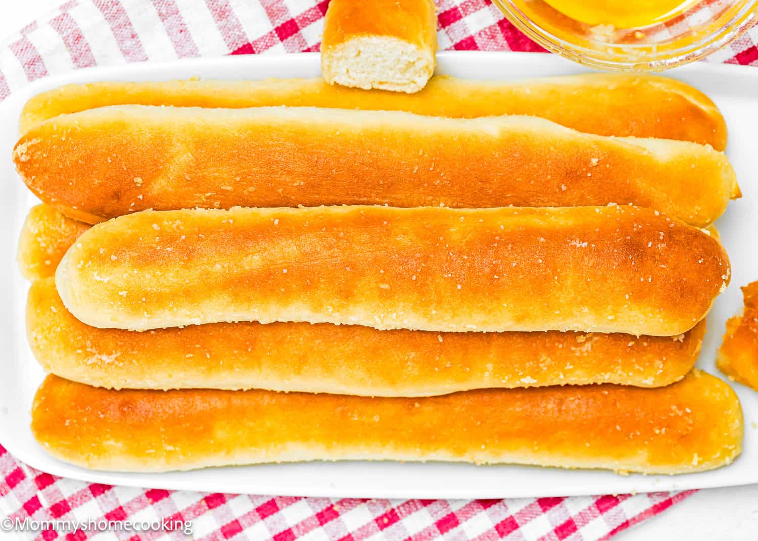 Golden breadsticks, quick and easy to make, are arranged on a white dish alongside a small bowl of honey. Perfect for those seeking egg-free and dairy-free options, these treats rest on a red and white checkered cloth.