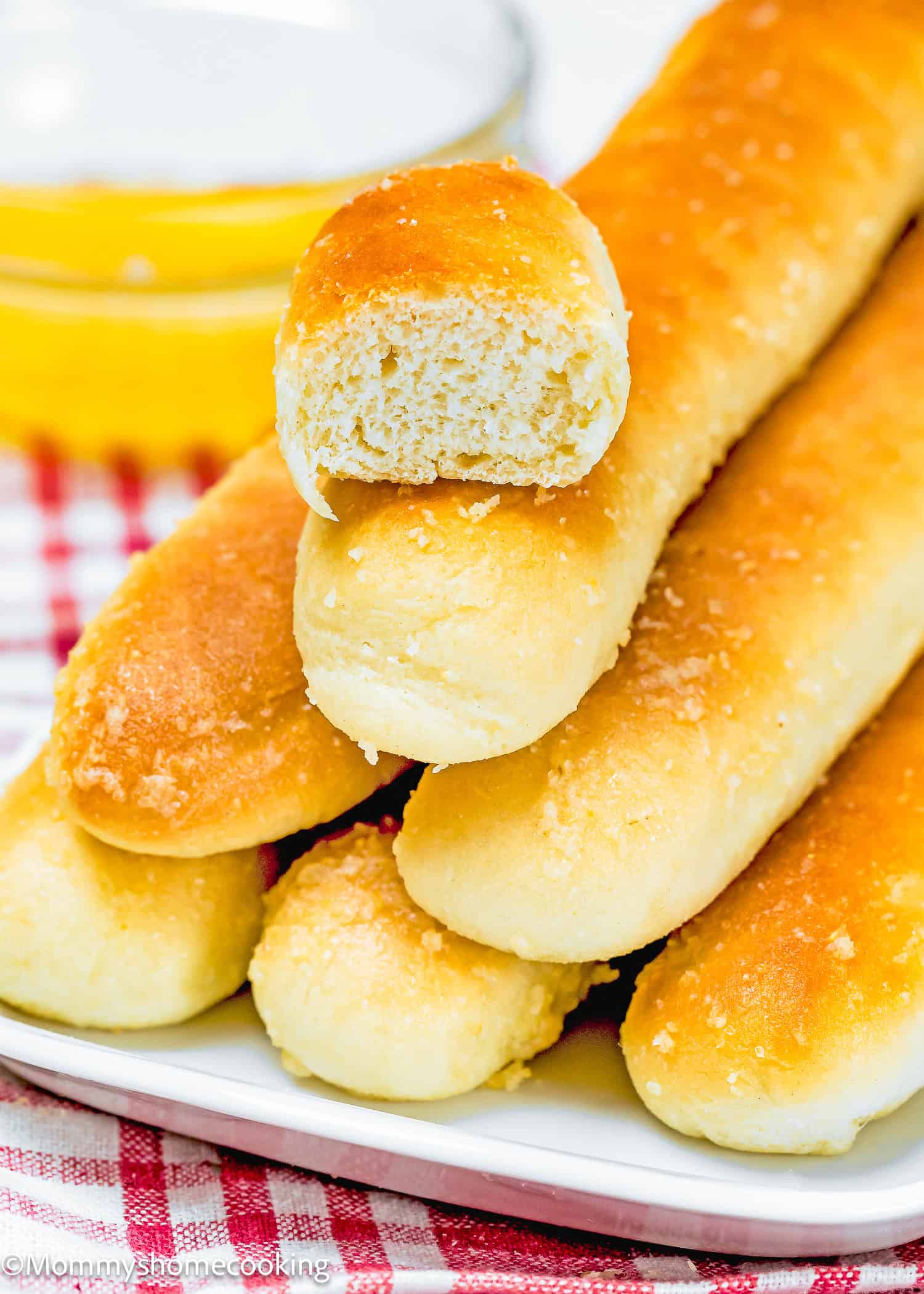 A pile of golden-brown, quick and easy breadsticks, delightfully egg-free and dairy-free, rests on a white plate. One has a bite taken out of it, hinting at its irresistible taste, while a small bowl of butter sits temptingly in the background.