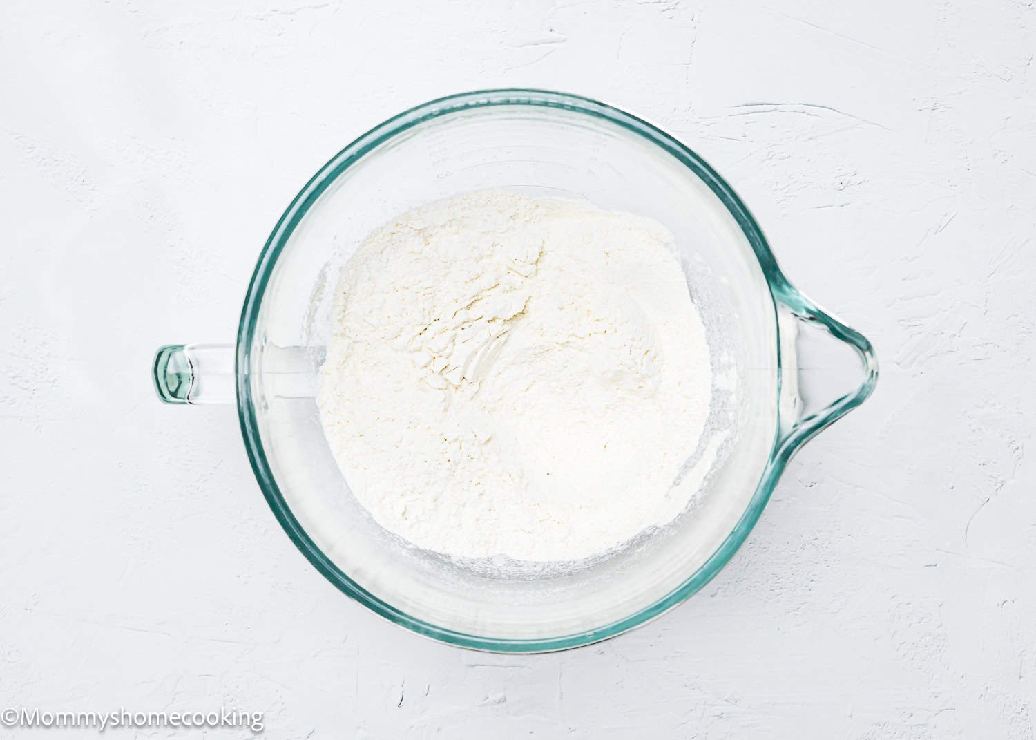 A clear glass bowl filled with white flour sits on a white textured surface, ready to create quick and easy breadsticks, perfectly egg-free and dairy-free.