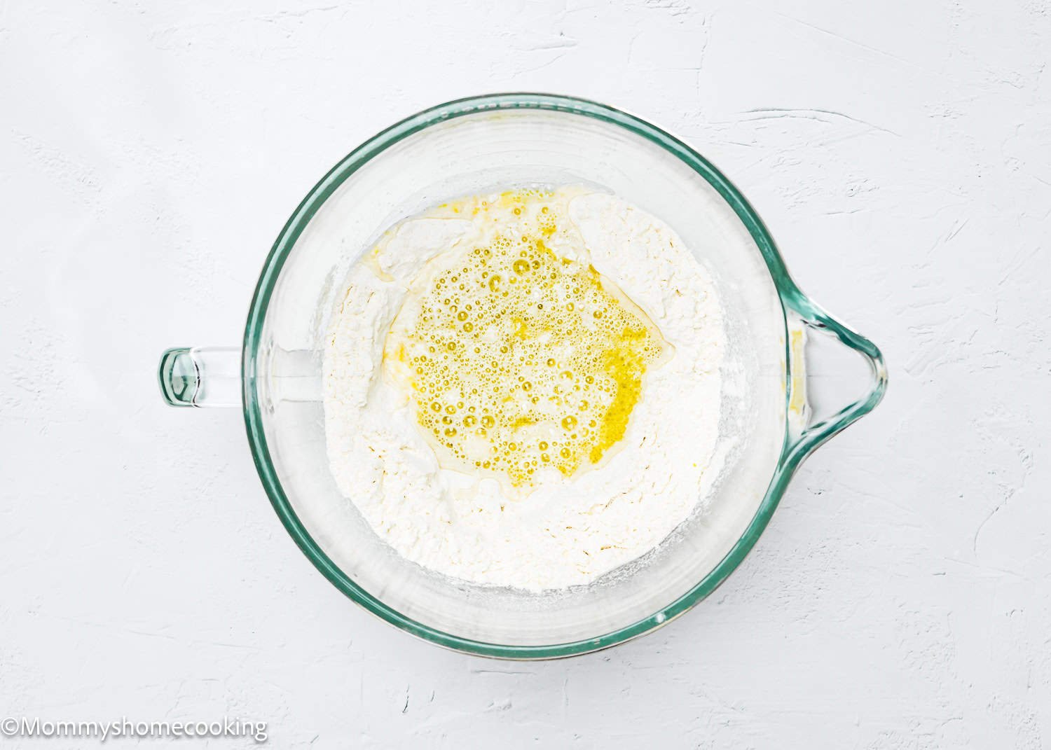 A glass mixing bowl containing flour and a yellow liquid sits on a white surface, poised for creating quick and easy breadsticks that are egg-free and dairy-free.