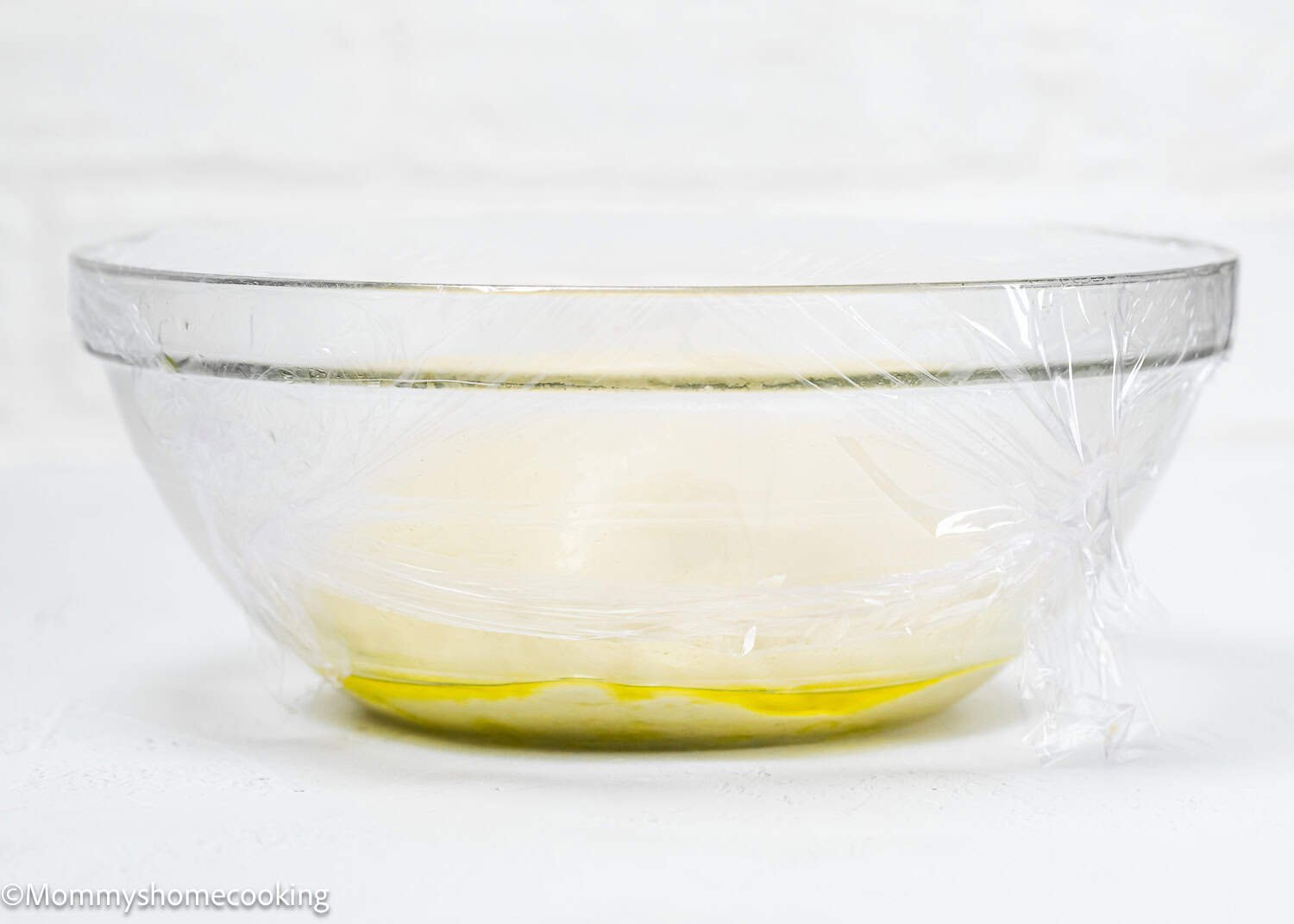 A glass bowl covered with plastic wrap holds the quick and easy breadstick dough, beginning to rise, with a hint of oil visible at the bottom. This egg-free and dairy-free recipe promises simplicity without sacrificing flavor.