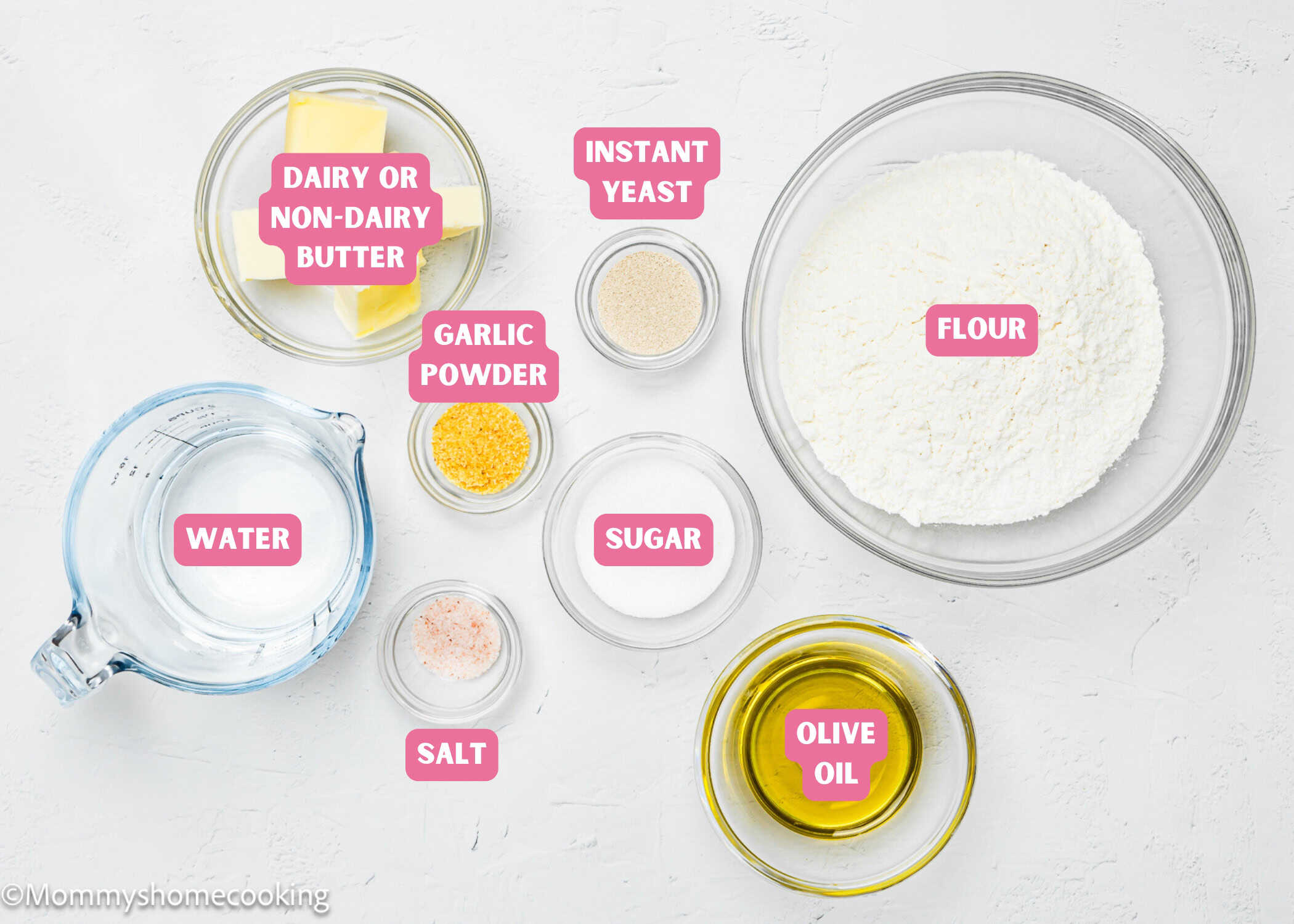 A neatly organized display of baking essentials on a white surface showcases flour, butter, water, yeast, garlic, sugar, salt, and oil in separate bowls—ideal for crafting quick and easy breadsticks that are both egg-free and dairy-free.