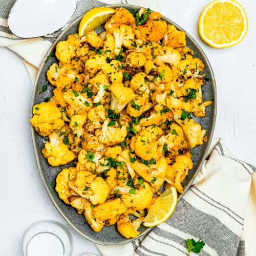 A quick and easy roasted cauliflower platter, seasoned with herbs and garnished with lemon wedges, rests on a striped cloth. Nearby are a spoon, a lemon half, small bowls of spices, and parsley leaves, inviting you to enjoy this effortless culinary delight.