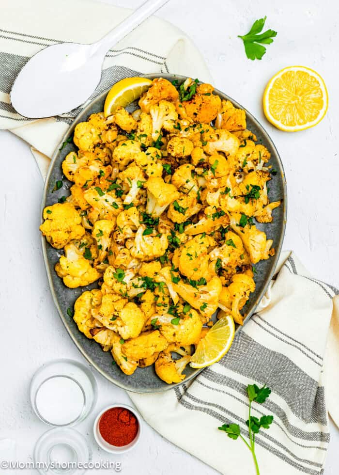 A quick and easy roasted cauliflower platter, seasoned with herbs and garnished with lemon wedges, rests on a striped cloth. Nearby are a spoon, a lemon half, small bowls of spices, and parsley leaves, inviting you to enjoy this effortless culinary delight.