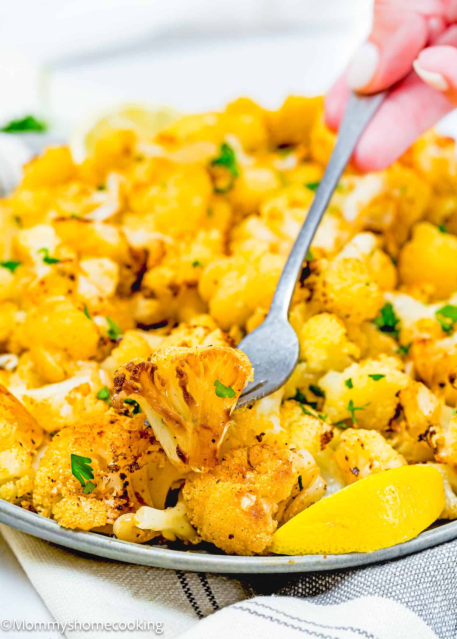 Close-up of quick and easy roasted cauliflower florets on a plate, with a fork lifting a piece. Slices of lemon are visible on the side, and the dish is garnished with herbs for an extra touch of flavor.