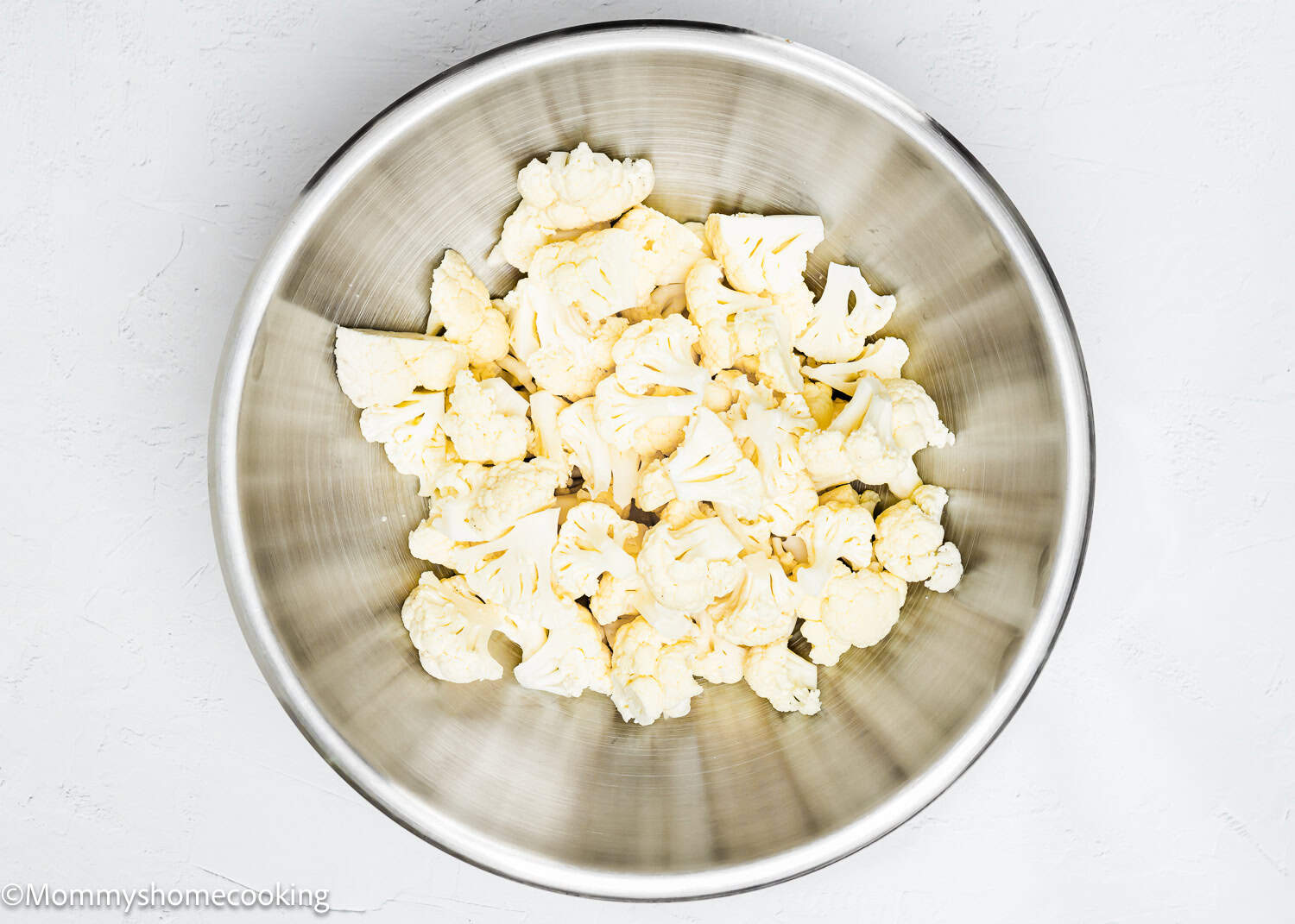 A stainless steel bowl filled with cauliflower florets rests on a light background.