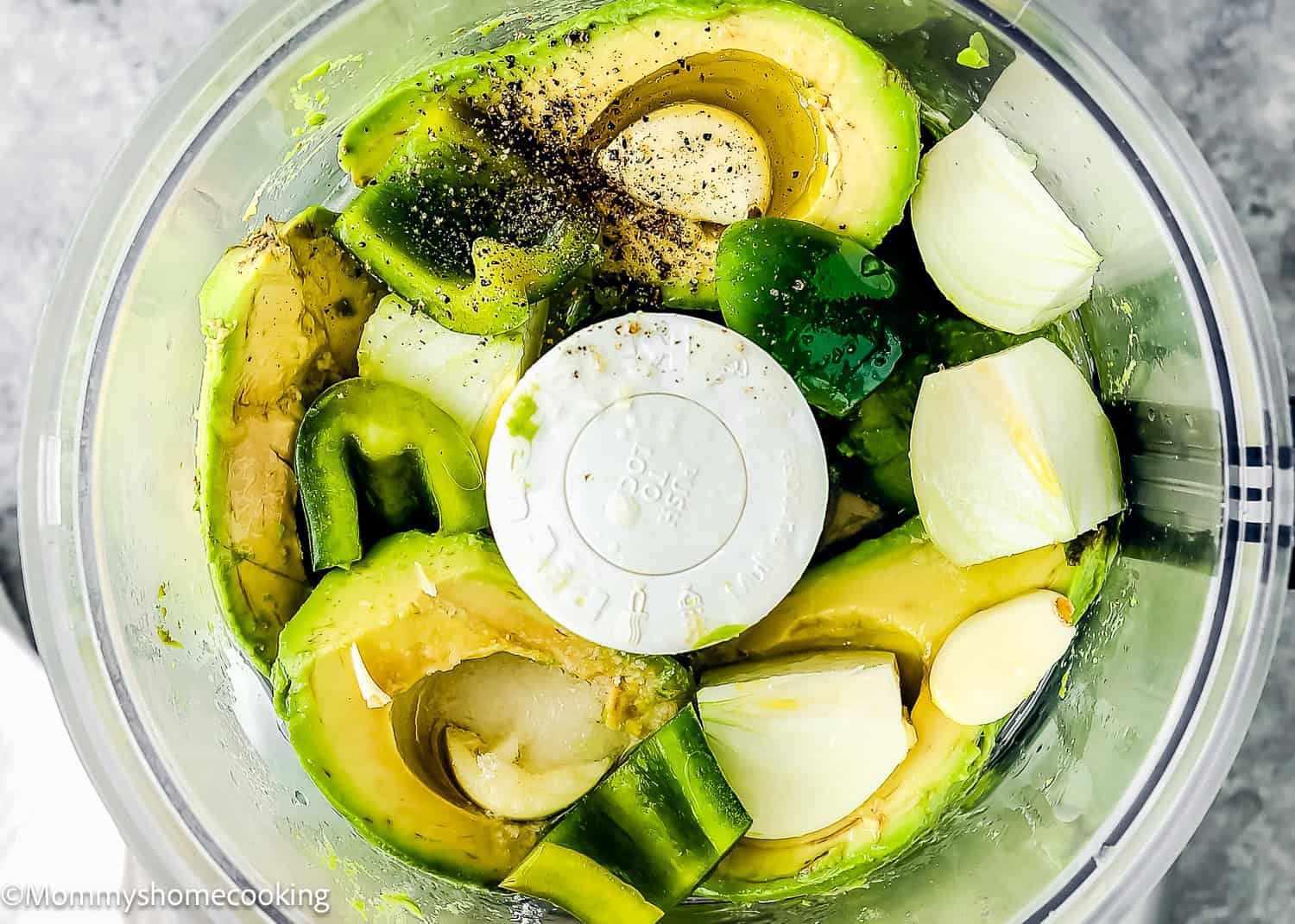 Venezuelan Guasacaca features avocado halves, onion chunks, garlic cloves, and green pepper pieces blended with black pepper in a food processor.