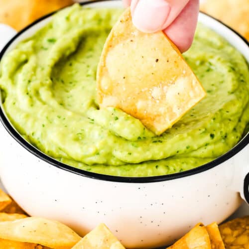 A hand dips a tortilla chip into a bowl of creamy Venezuelan Guasacaca, with additional chips scattered nearby.