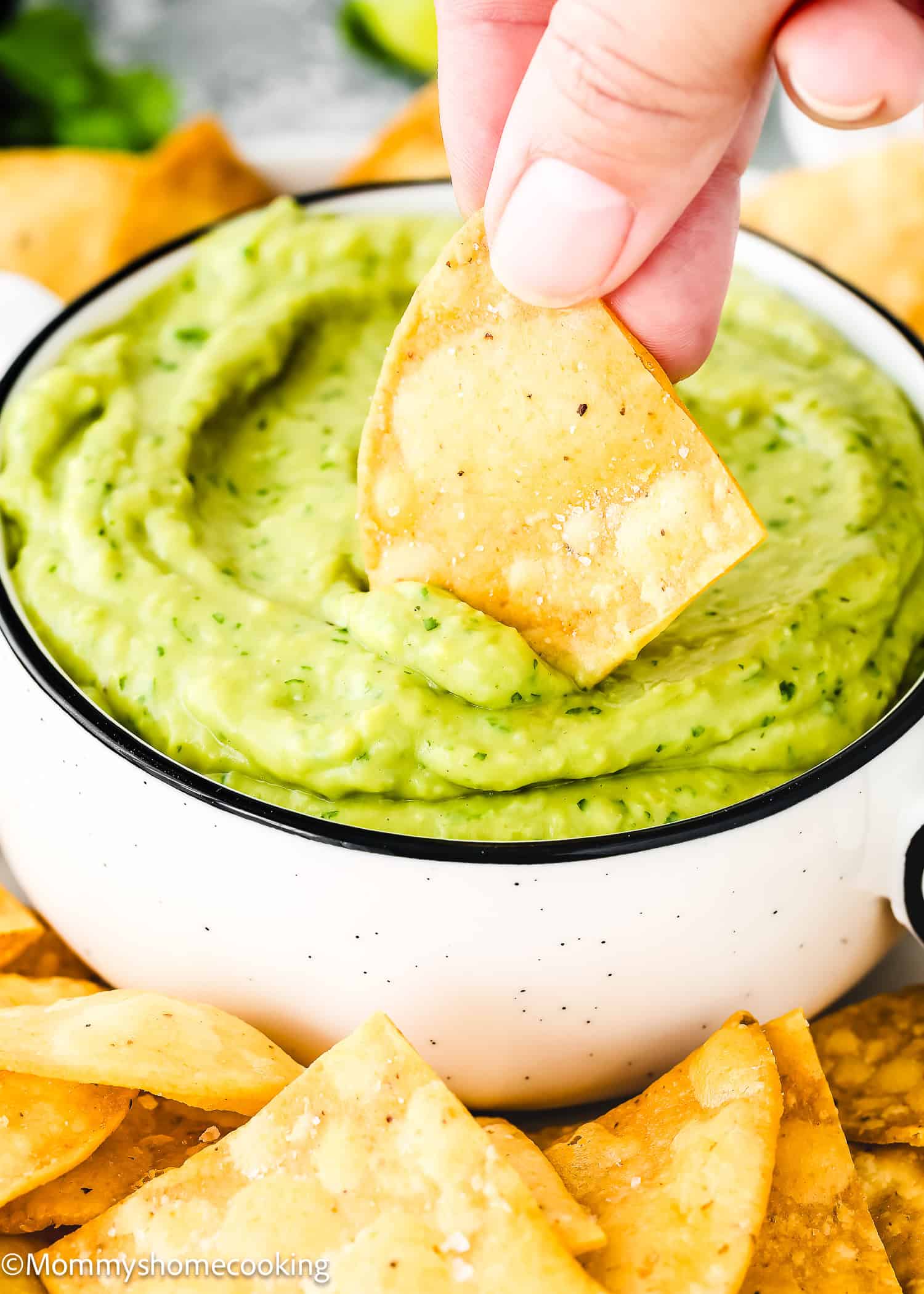 A hand dips a tortilla chip into a bowl of creamy Venezuelan Guasacaca, with additional chips scattered nearby.
