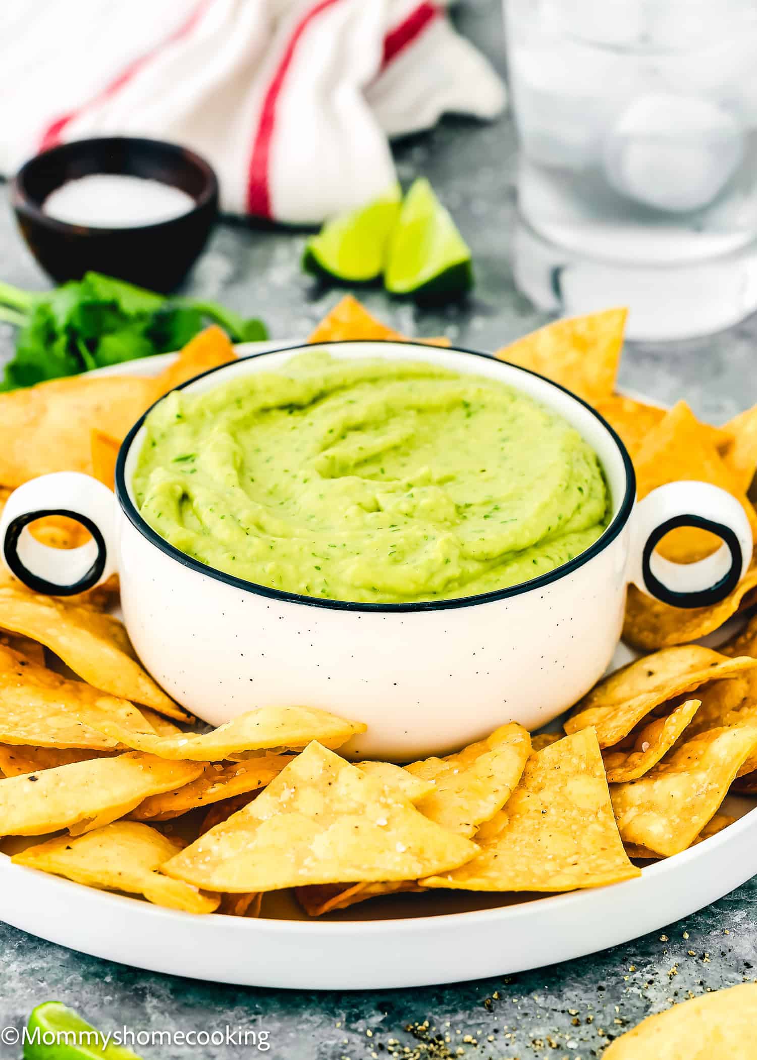 A bowl of Venezuelan Guasacaca surrounded by tortilla chips on a white plate, with lime wedges and a drink in the background.
