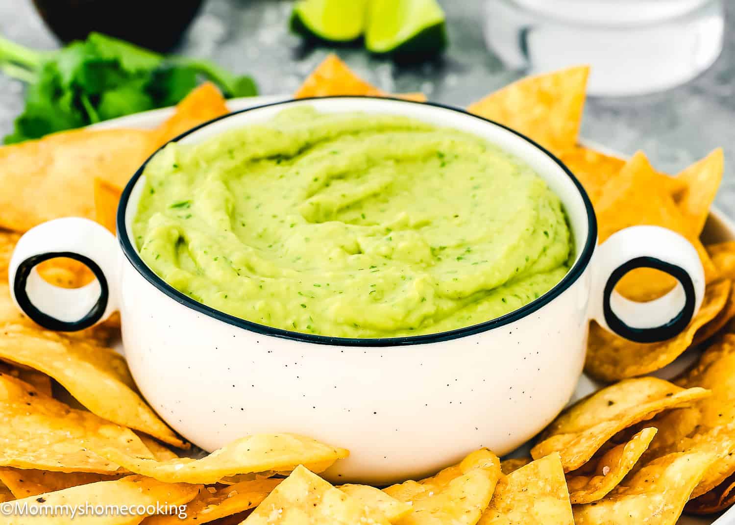 A bowl of Venezuelan Guasacaca surrounded by tortilla chips on a plate. Lime wedges and cilantro are blurred in the background.