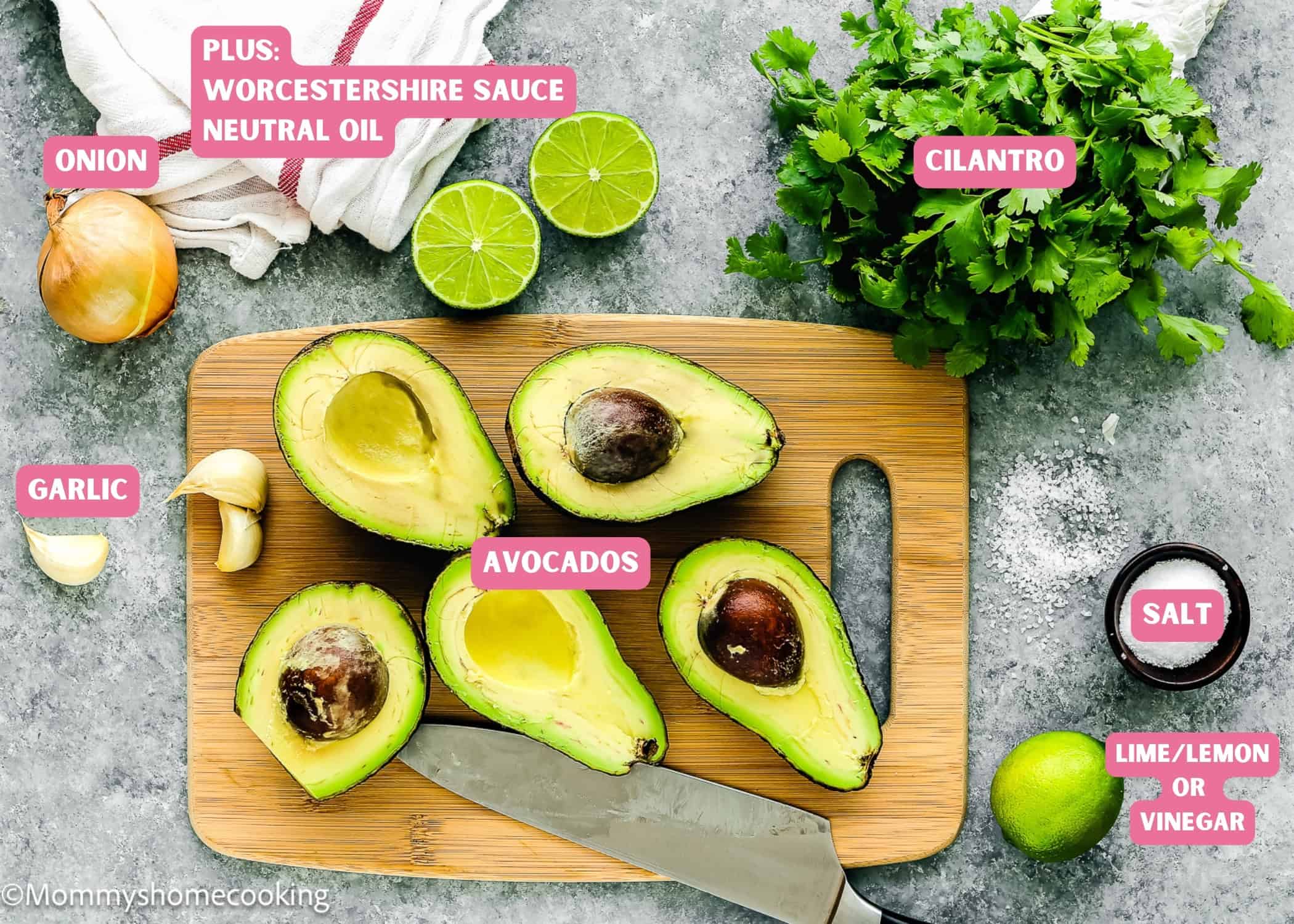 Ingredients for making guasacaca on a wooden board: avocados, onion, garlic, lime, cilantro, and salt. A hint of Worcestershire sauce and neutral oil nods to Venezuelan Guasacaca. A knife rests beside the avocados.
