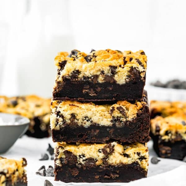 A stack of three chocolate chip cookie brownies, known as the best eggless brookies, sits on a white surface with scattered chocolate chips and a bottle of milk in the background.