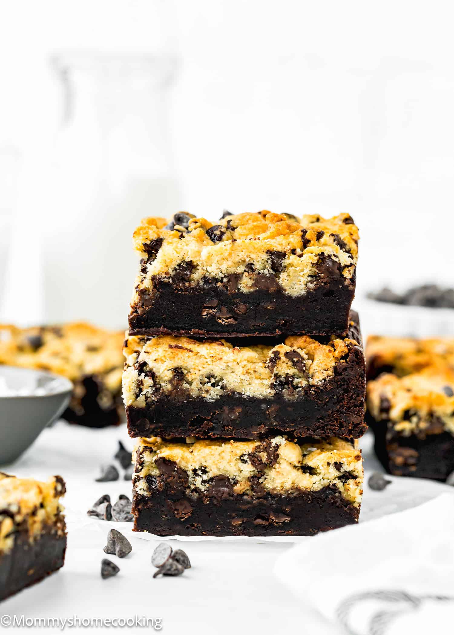 A stack of three chocolate chip cookie brownies, known as the best eggless brookies, sits on a white surface with scattered chocolate chips and a bottle of milk in the background.