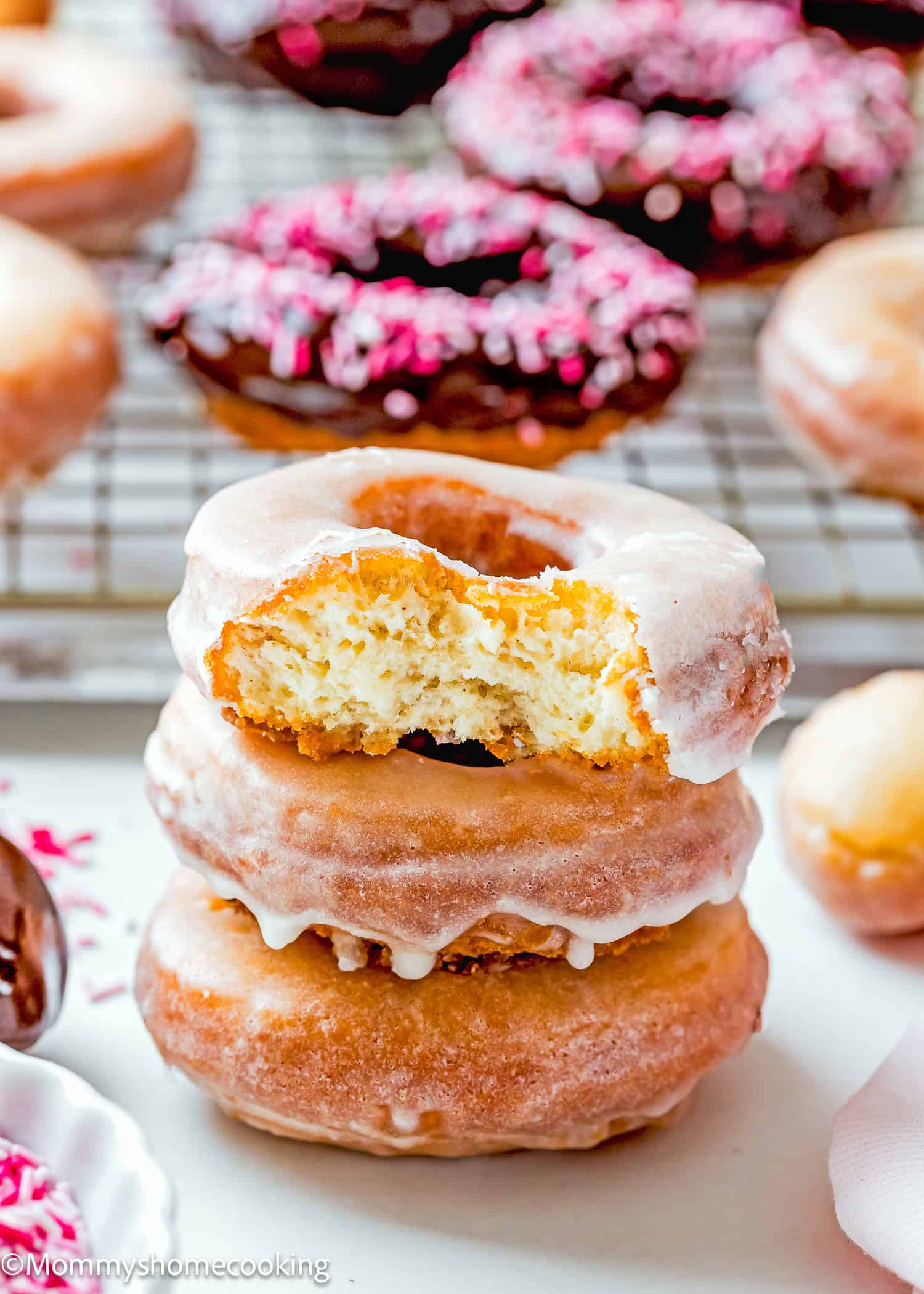 A stack of three glazed donuts, reminiscent of the best eggless cake donuts—quick and easy to make—with a bite taken out of the top one. In the background, more donuts, some adorned with pink sprinkles, rest on a wire rack.