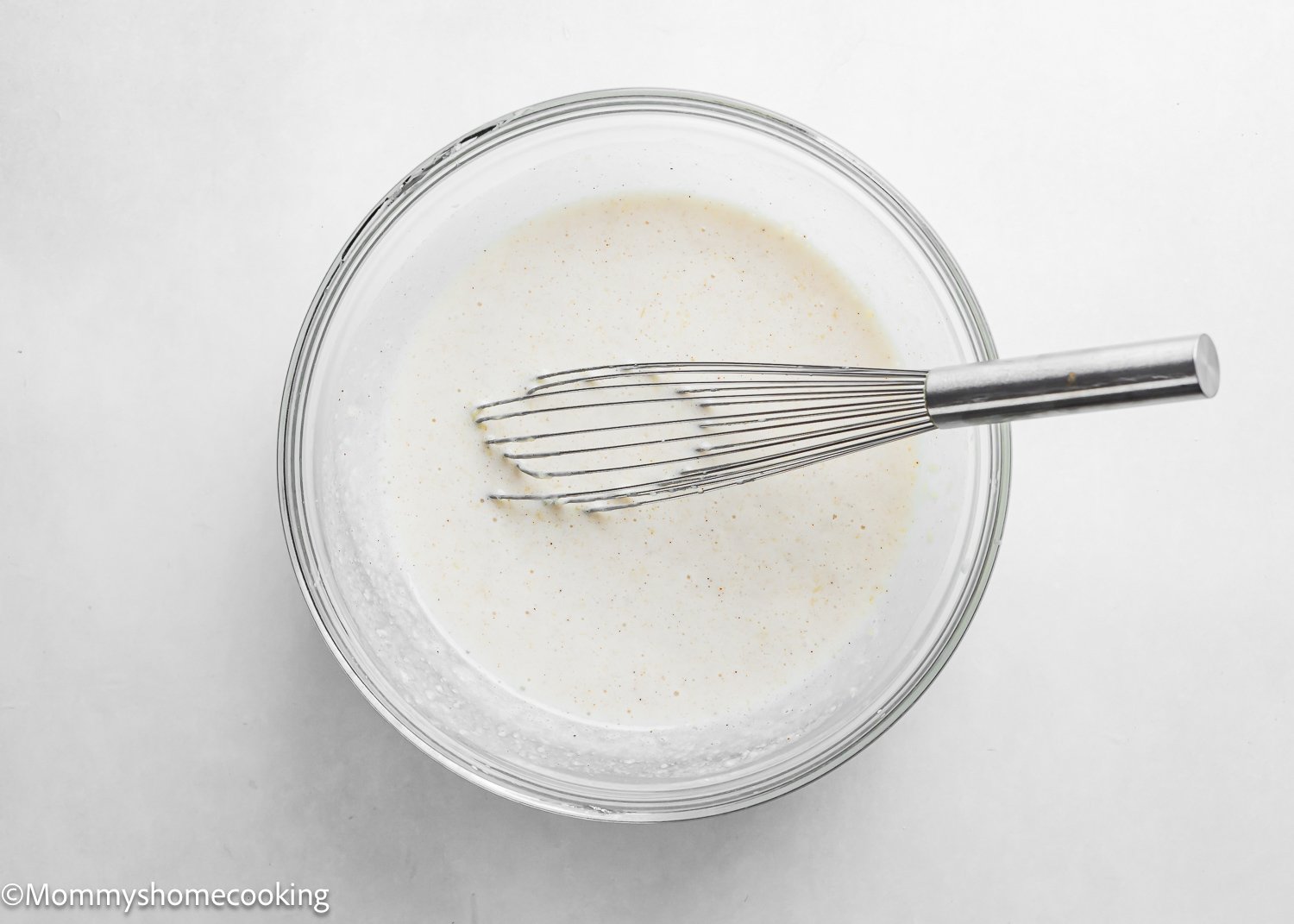 A mixing bowl with a metal whisk rests in a creamy white liquid, possibly the batter for Best Eggless Cake Donuts: Quick & Easy, on a pristine white surface.