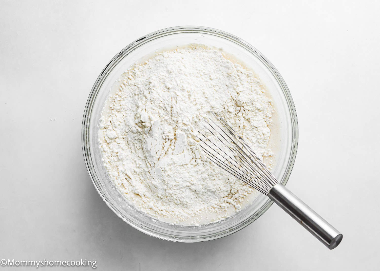 A mixing bowl containing flour with a whisk placed on top, set against a plain white background—perfect for preparing the best eggless cake donuts, quick & easy.