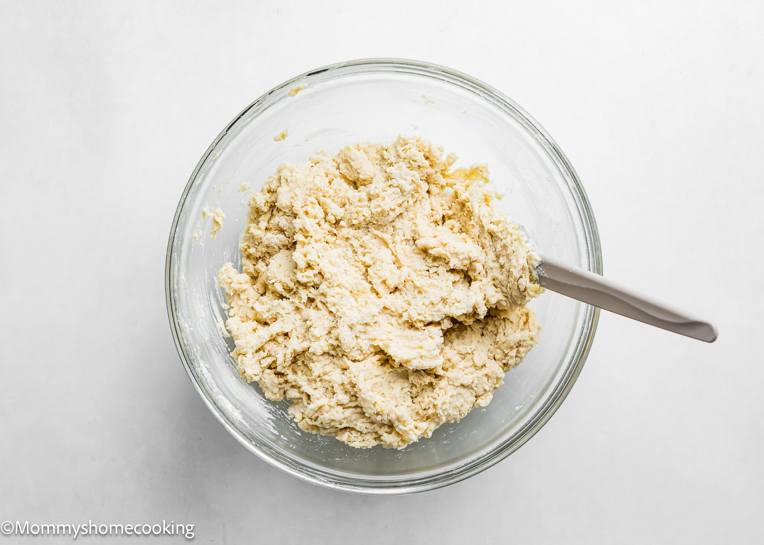 A clear glass bowl filled with thick, lumpy dough for the Best Eggless Cake Donuts (Quick & Easy). A spatula rests in the bowl, ready to mix. The background is a plain white surface.