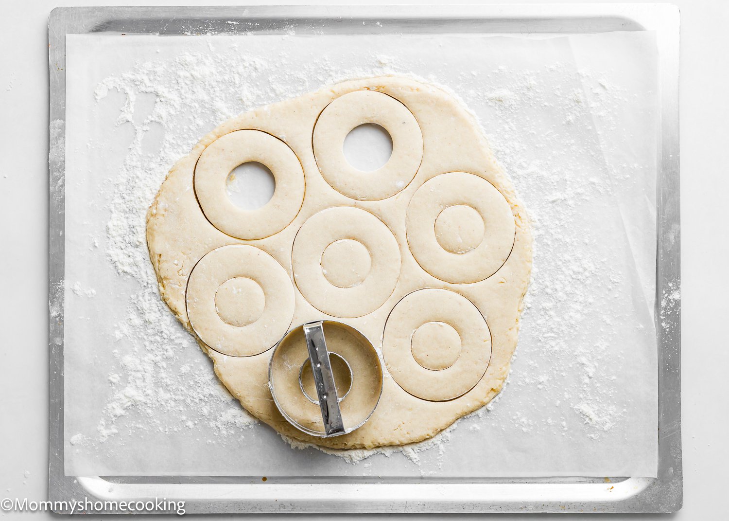 Dough rolled out on a baking sheet with round donut shapes cut out, showcasing the best eggless cake donuts. A round cutter lies on the dough, making it quick and easy to create delicious treats.