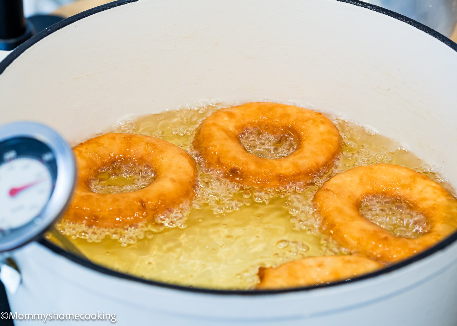 Four doughnuts frying in oil in a white pot, bubbles forming around them. A thermometer is clipped to the pot's side, ensuring the perfect temperature for these quick and easy best eggless cake donuts.