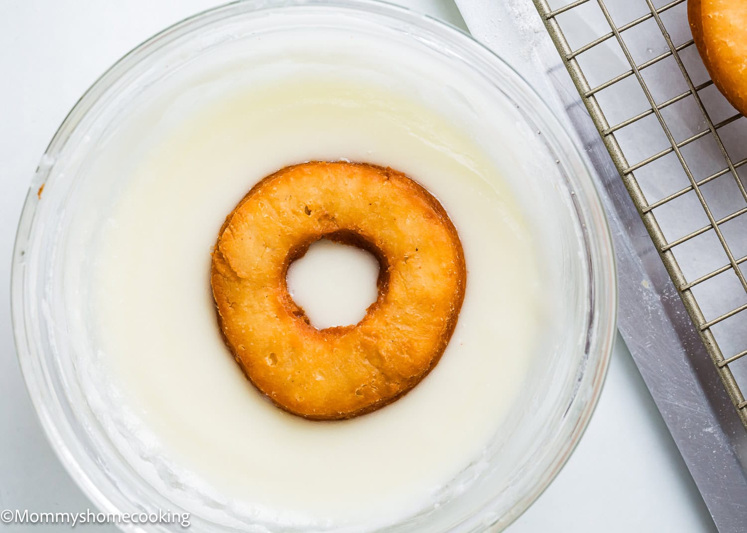 A donut is partially dipped in white glaze in a glass bowl, capturing the essence of Best Eggless Cake Donuts. Another donut sits on a cooling rack nearby, showcasing how quick and easy it is to create these delightful treats.